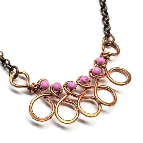 The Copper Wire Wrapped Sculpted Pink Gemstone Necklace by Alexa Martha Designs features five large copper wire-wrapped loops on a dark chain, each topped with a pink gemstone for an artistic pendant look.