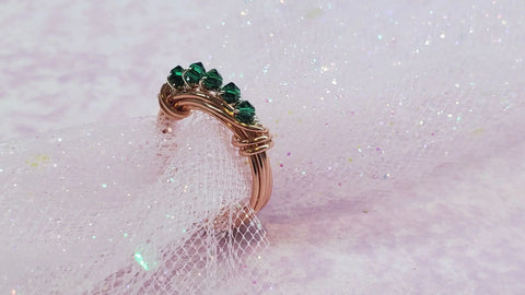 The Wire Wrapped Copper Crystal Birthstone Bling Ring by Alexa Martha Designs, featuring a row of green crystals, is displayed on white glittery tulle with a soft pastel background.