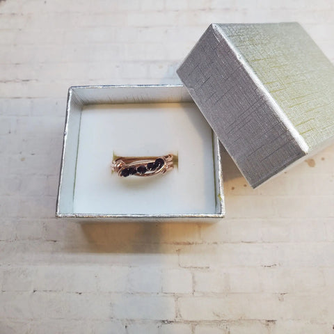 The Alexa Martha Designs Wire Wrapped Copper Crystal Birthstone Bling Ring with dark gemstones is shown in a white and silver ring box with a partially open lid, placed on a light-colored textured surface.