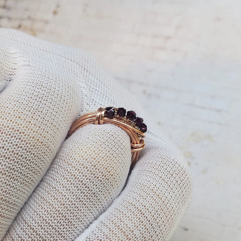 A close-up of a white-gloved hand holding the Wire Wrapped Copper Crystal Birthstone Bling Ring by Alexa Martha Designs, featuring gold wire and a row of small dark red stones against a softly blurred background.