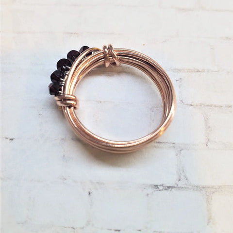 The Wire Wrapped Copper Crystal Birthstone Bling Ring by Alexa Martha Designs features entwined rose gold bands and a cluster of dark Swarovski crystals, displayed on a light, textured surface.