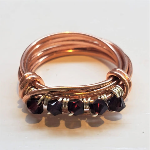 The Wire Wrapped Copper Crystal Birthstone Bling Ring by Alexa Martha Designs features multiple bands accented with dark red beads and sparkling Swarovski crystals, all secured with gold wire along the front.
