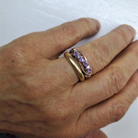 A hand wears a plain gold band on the ring finger and Alexa Martha Designs Wire Wrapped Copper Crystal Birthstone Bling Ring, sparkling with five purple stones, on the middle finger against a white background.