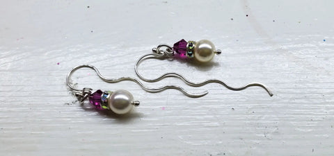 The Alexa Martha Designs Ergonomic Crystal Pearl Wavy Ear Threaders feature handmade Swarovski crystals, white pearl beads, purple accents, and sparkling spacers on silver hooks, displayed on a white wooden surface.