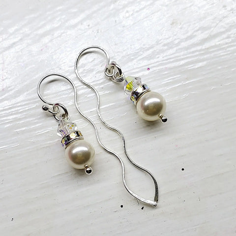 The Ergonomic Crystal Pearl Wavy Ear Threaders by Alexa Martha Designs, featuring wavy hooks, white pearls, and clear crystal beads, are displayed on a textured white surface.