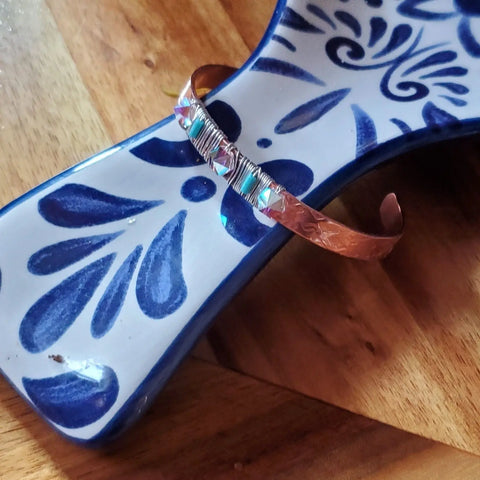 The Crystal Spike Bead Copper Cuff by Alexa Martha Designs, featuring copper, turquoise beads, and silver wire wrapping, is displayed on a blue and white floral ceramic piece atop a wooden surface.
