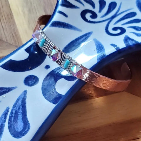 The Crystal Spike Bead Copper Cuff by Alexa Martha Designs is a punk-style copper bangle engraved with patterns, wrapped in silver wire, and decorated with turquoise and pink beads, displayed on a blue and white ceramic piece.