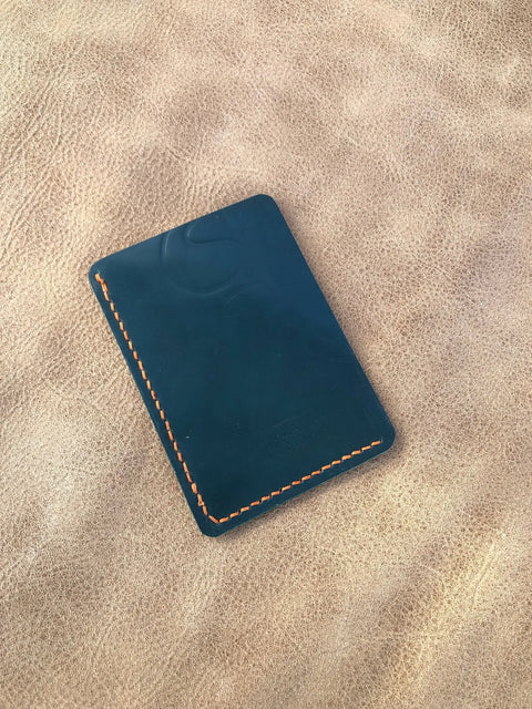 The Sully + Proper Supply Deep Sea Teal Card Wallet, featuring orange stitching, rests on textured tan leather, highlighting this handcrafted minimalist wallet made in the USA.