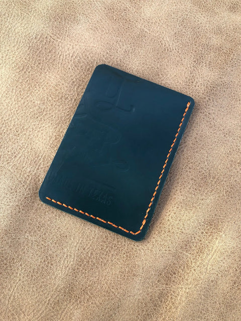 A Deep Sea Teal Card Wallet by Sully + Proper Supply rests on a textured tan surface. This slim, minimalist wallet showcases embossed text, including MADE IN TEXAS, on the front.