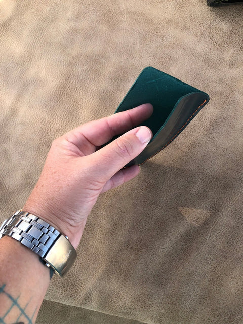 A person with a tattooed wrist and metal watch holds an open Sully + Proper Supply Deep Sea Teal Card Wallet, handcrafted in the USA, against a textured brown surface.