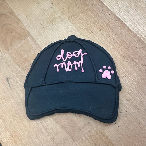 The Dogtown Bakery Dog Mom Hat Cookie is decorated like a black baseball cap with “dog mom” in pink icing and a pink paw print—ideal for personalized gifts or fans of artisan pet-themed treats.