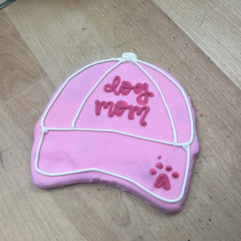 Dogtown Bakery’s Dog Mom Hat Cookie is a pink, baseball cap-shaped treat with white trim, “dog mom” in red icing, and a small paw print detail—an artisanal pet-themed gift perfect for any dog lover.