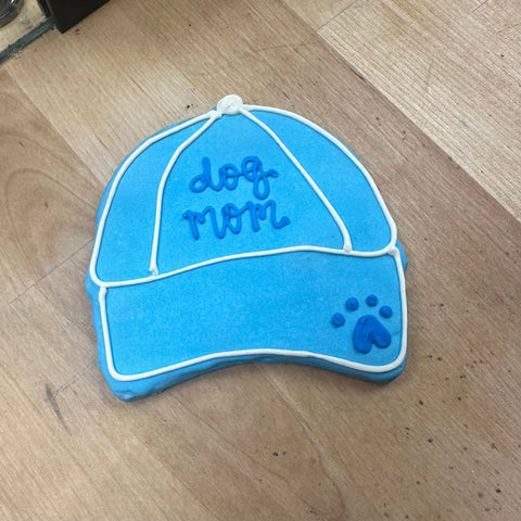 The Dog Mom Hat Cookie by Dogtown Bakery is a blue, hat-shaped artisan cookie with white and dark blue icing and a paw print heart on the brim—an adorable, pet-themed gift displayed on a light wood surface.