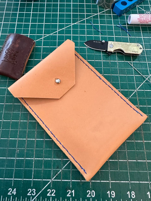 A Sully + Proper Supply Utility Pouch, crafted from tan English Bridle Leather with blue stitching and a snap closure, sits on a green cutting mat among a folding knife, brown leather item, and assorted crafting tools.