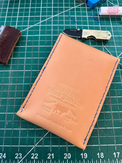 A tan English Bridle Leather wallet with blue stitching and embossed text sits on a green cutting mat. Nearby is a Sully + Proper Supply Utility Pouch, made in the USA from brown leather, plus crafting tools and a small folding knife with keychain.