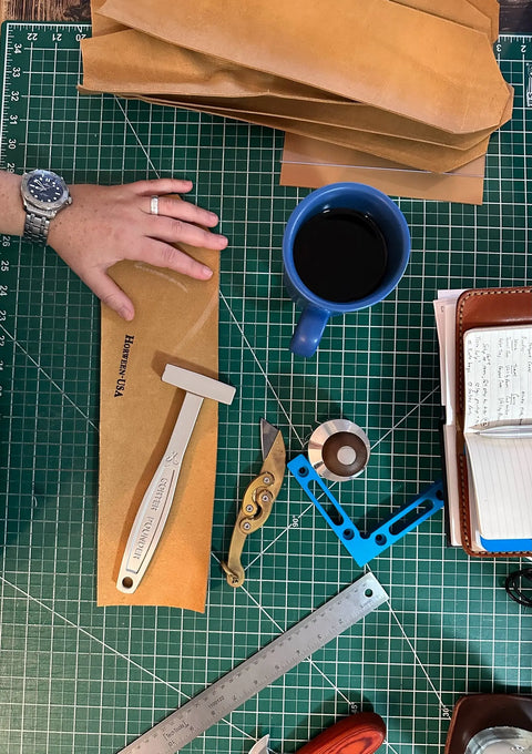 A hand rests on the Sully + Proper Supply Utility Pouch made from tan English Bridle Leather, placed beside a hammer, knife, blue coffee mug, ruler, tape, stitching tool, and notebook on a green cutting mat.