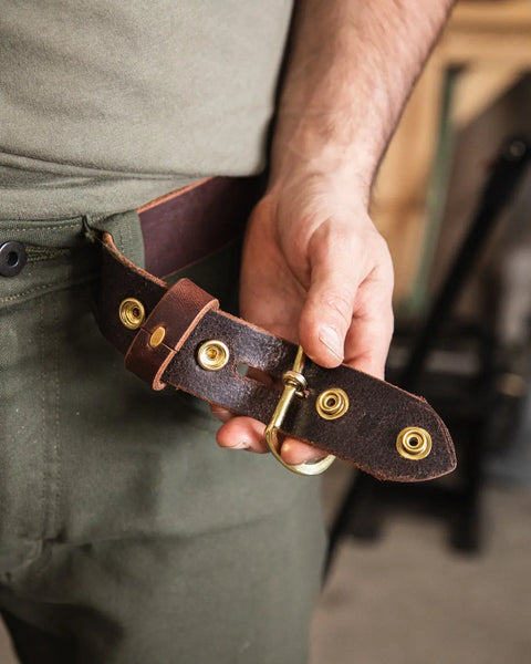 A person in green pants holds the Homestead Brand Essential Leather Belt No. 1508, featuring gold metal hardware with a solid brass buckle and rivets, in their hand.