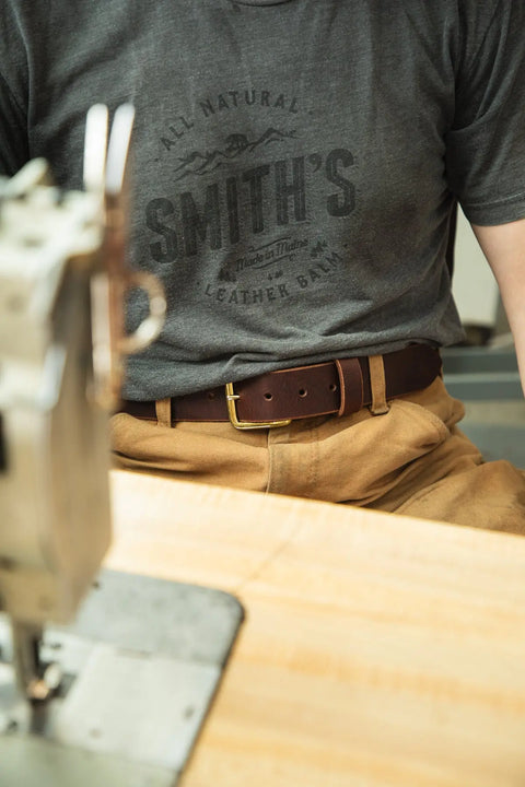 A person in a dark grey t-shirt and light brown pants sits at a wooden table by a sewing machine, perhaps crafting the Essential Leather Belt No. 1508 by Homestead Brand, featuring a solid brass buckle and made in the U.S.A.