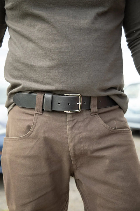 Shown from the waist down, a person wears brown pants with a dark olive long-sleeve shirt tucked in and an Essential Leather Belt No. 1508 by Homestead Brand, featuring black leather and a solid brass buckle.