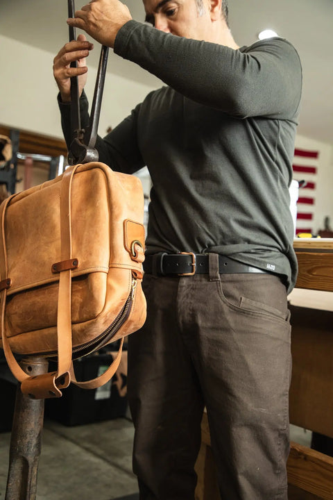 Wearing a dark long-sleeve shirt and brown pants, a person crafts the Essential Leather Belt No. 1508 by Homestead Brand in a workshop. An American flag in the background highlights its proudly Made in the U.S.A. craftsmanship.