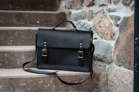 The Urban Southern Executive Briefcase, crafted from black full-grain leather with gold buckles and a shoulder strap, rests on a stone staircase against a rugged stone wall.
