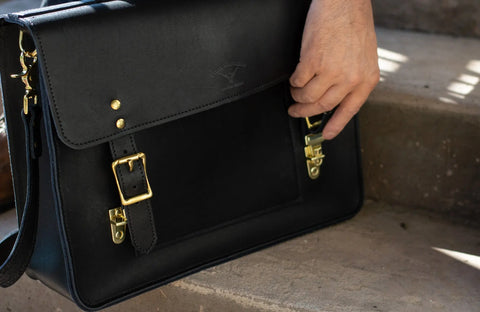 A hand opens the front buckle of the Urban Southern Executive Briefcase, a black full-grain leather satchel with gold hardware, resting on stone steps in sunlight.