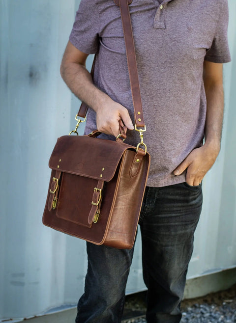 Wearing a short-sleeved purple shirt and black jeans, a person holds the Urban Southern Executive Briefcase, featuring full-grain leather and gold buckles, while standing before a light blue metal wall.
