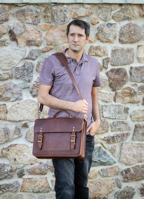 A man in a purple polo and black jeans stands before a stone wall, looking aside with one hand in his pocket while carrying the Urban Southern Executive Briefcase, a professional full-grain leather shoulder bag.