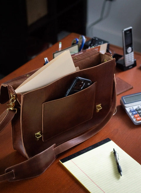 An Urban Southern Executive Briefcase, crafted from full-grain leather and holding papers and a phone, rests on a desk alongside a yellow notepad, pen, calculator, office phone, and pens in a holder.