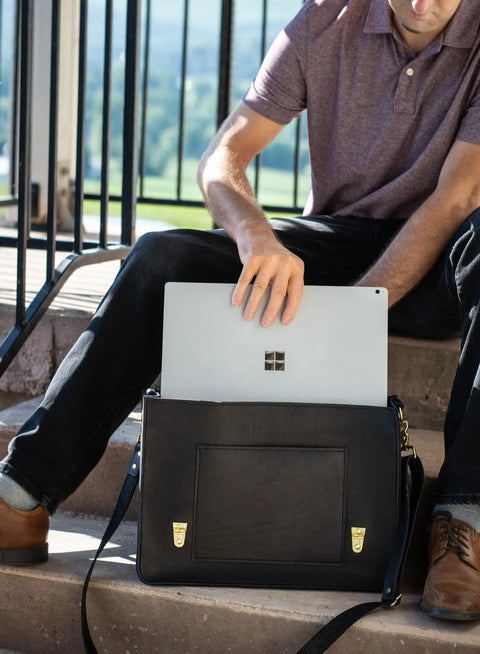 A person on outdoor steps in a short-sleeve shirt and dark pants puts a Microsoft Surface laptop into an Urban Southern Executive Briefcase made from full-grain leather.