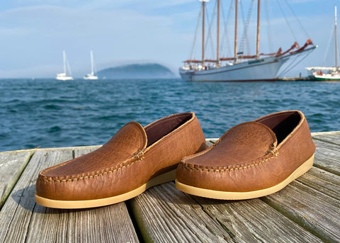 A pair of BBH Daily Moc brown full-grain leather loafers with tan soles rest on a wooden dock by the water, sailboats and a large ship anchor in the background beneath a partly cloudy sky.