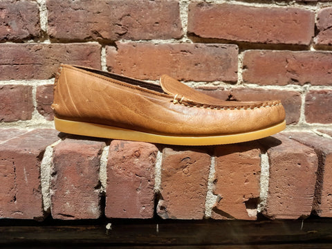 The BBH Daily Moc, a tan full-grain leather loafer with a light rubber sole, is shown on a sunlit brick ledge against a red brick wall.