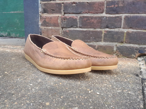 A pair of BBH Daily Moc brown full-grain leather Venetian-style moccasins with light tan soles is displayed on rough concrete against a weathered brick wall.
