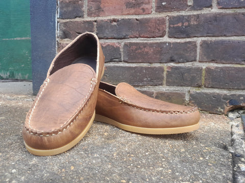 The BBH Daily Moc is a handcrafted, brown full-grain leather Venetian-style loafer with tan soles, made in the USA and shown outdoors on rough concrete with a brick wall and green door in the background.