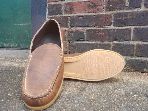 The BBH Daily Moc, a pair of brown Venetian-style moccasin loafers handcrafted in the USA from full-grain leather, is shown outdoors by a brick wall—one upright, the other on its side to display the tan, textured sole.