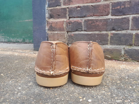 A pair of BBH Daily Moc brown full-grain leather moccasins with beige soles, handcrafted in the USA, rest on a rough concrete surface with heels facing the camera. A brick wall and green door are visible in the background.