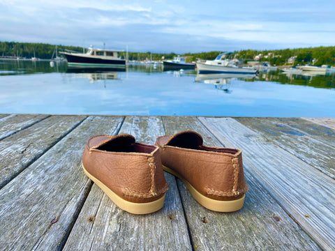 The BBH Daily Moc, crafted from brown full-grain leather, rests on a wooden dock overlooking calm water and boats beneath a partly cloudy sky.