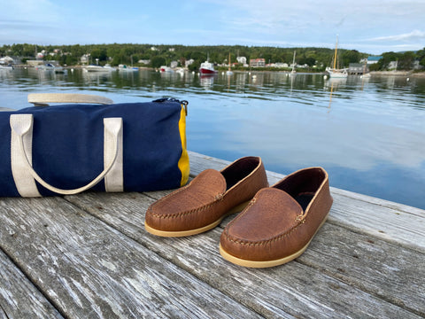 The BBH Daily Moc brown full-grain leather loafers and a blue-and-yellow duffel bag sit on a wooden dock by calm water, with boats and a green shoreline in the background beneath a partly cloudy sky.