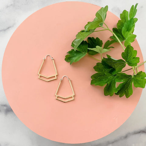 The GOLD CLEO HOOPS by Sarah Cecelia are displayed with a green leafy sprig on a round peach-colored surface, set against a marble background.