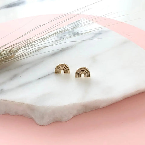 Sarah Cecelias GOLD ARCO STUDS are small gold rainbow-shaped earrings, pictured on white marble with dried grass and a pastel pink backdrop—perfect for adding a subtle touch of LGBTQIA pride to any look.