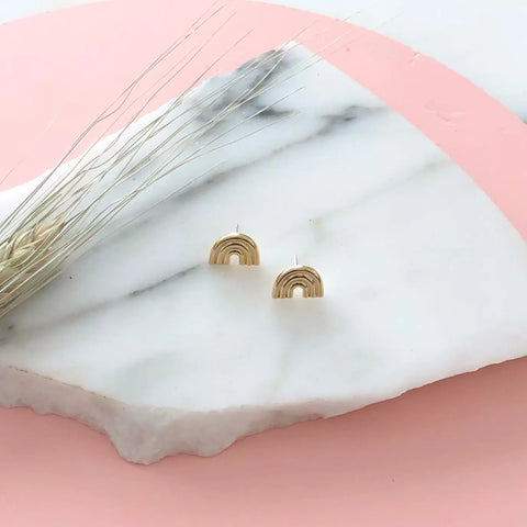 A pair of small GOLD ARCO STUDS by Sarah Cecelia sits on a white marble slab with soft pink edges, accented by dried wheat on the left. These gold rainbow earrings offer a subtle nod to LGBTQIA pride.