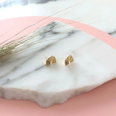 GOLD ARCO STUDS by Sarah Cecelia—elegant rainbow-shaped earrings—rest on a round white marble slab against a pink backdrop with dried grass. Celebrate LGBTQIA pride with this stylish jewelry statement.