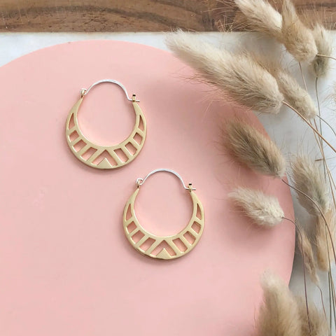 The Sarah Cecelia GOLD SPECTRA HOOPS, featuring geometric gold cut-out designs, are shown on a pink surface beside beige pampas grass.