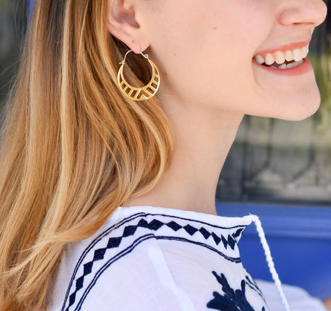A blonde woman smiles in a white top with navy blue embroidery, wearing large GOLD SPECTRA HOOPS by Sarah Cecelia. The background is softly blurred.