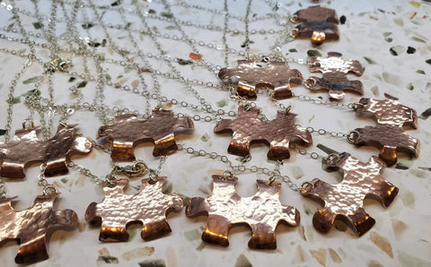 The Handmade Hammered Copper Puzzle Piece Necklace by Alexa Martha Designs features a unique copper pendant on a silver chain, beautifully displayed on a textured white and stone-speckled surface. Perfect as handmade autism jewelry.