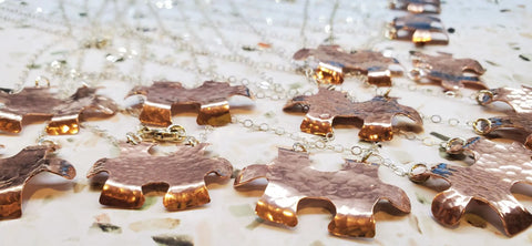 Handmade Hammered Copper Puzzle Piece Necklaces by Alexa Martha Designs, featuring textured, shiny pendants on silver chains, are displayed on a speckled surface. These autism jewelry pieces beautifully reflect light.