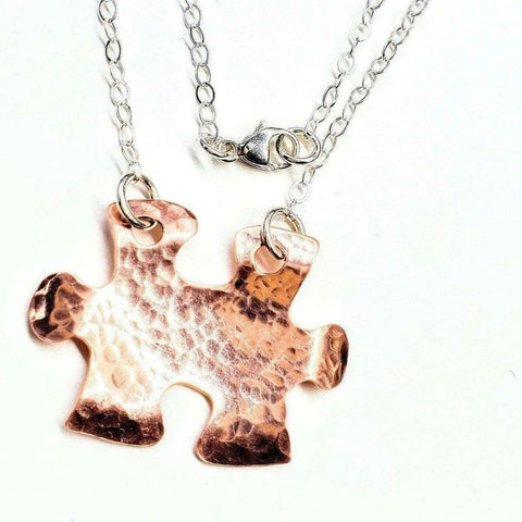 The Handmade Hammered Copper Puzzle Piece Necklace by Alexa Martha Designs features a textured copper pendant on a silver chain, shown against a white background. This unique autism jewelry is handmade and attached with metal rings.
