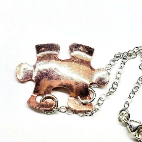 The Handmade Hammered Copper Puzzle Piece Necklace by Alexa Martha Designs, featuring a copper puzzle pendant on a silver chain, is displayed against a white background.