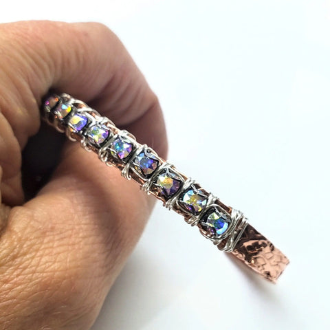 A hand holds a Set of Glamorously Wrapped Hammered Rhinestone Crystal Bling Bangles by Alexa Martha Designs, featuring iridescent gemstones nestled in silver wire with a hammered shine, echoing stylish handcrafted cuff trends.
