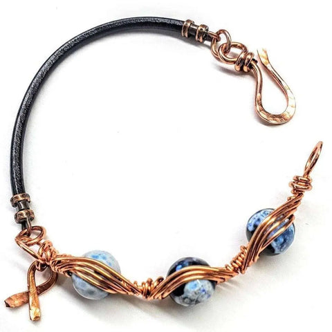 The Herringbone Wire Wrap Child Abuse Prevention Half Cuff Ribbon Bracelet by Alexa Martha Designs features a black leather band, blue marbled beads, copper wire wrap, hook clasp, and a copper awareness ribbon charm.